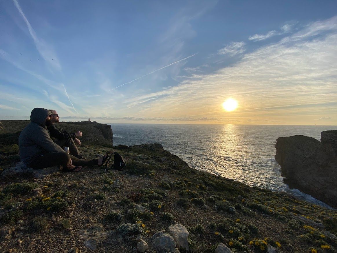Two people sit looking over the cliffs to the ocean while the sun sets